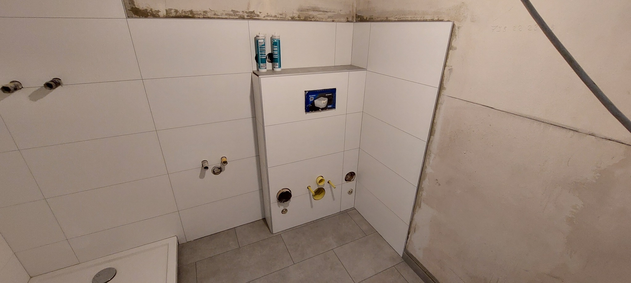 Wnętrze łazienki w trakcie wykańczania: białe, kwadratowe płytki na ścianach, zabudowa stelaża podtynkowego WC z widocznymi przyłączami hydraulicznymi i rurami, surowa ściana w tle.