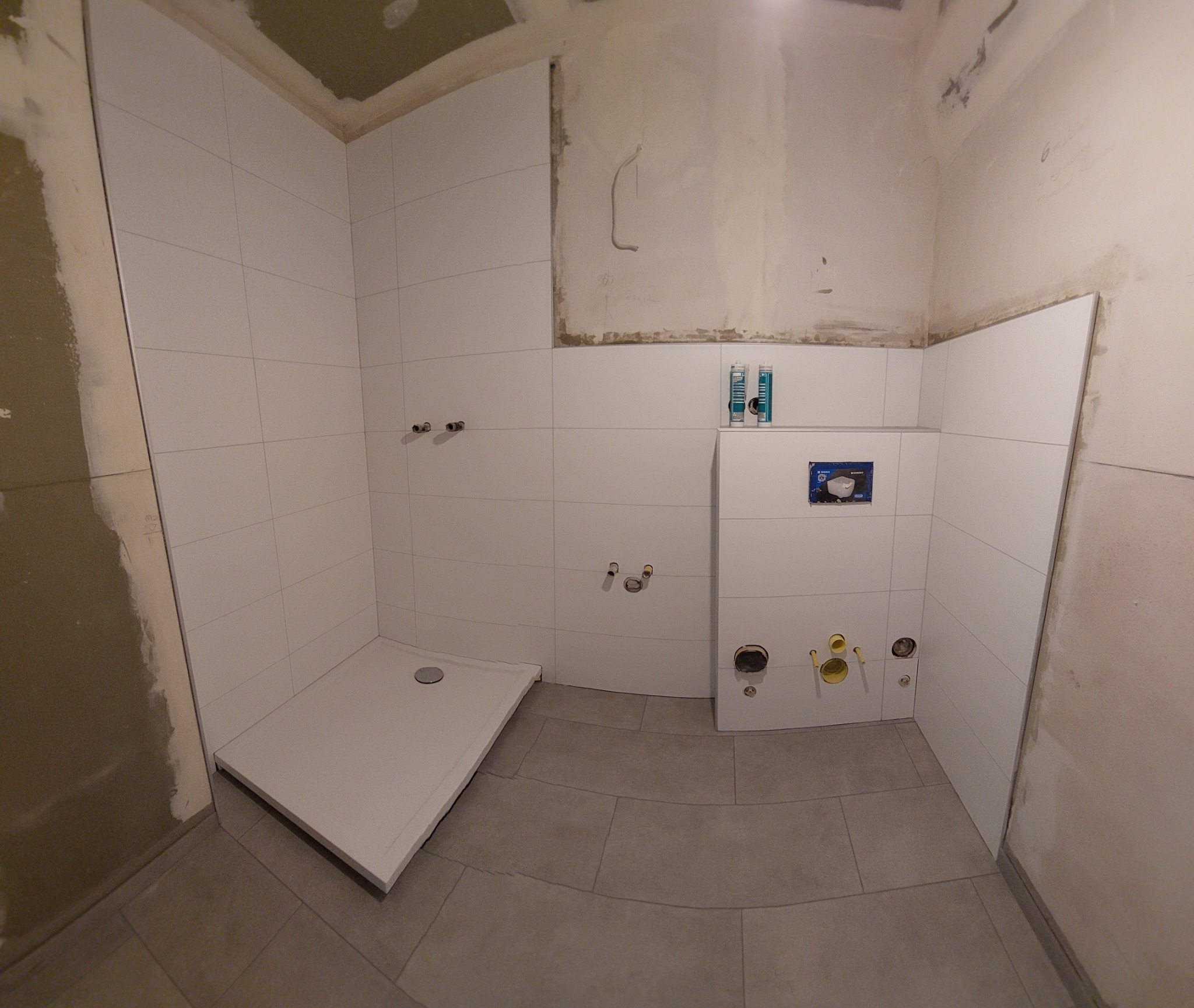 Wnętrze łazienki w trakcie remontu w Salzgitter, widoczny brodzik, białe płytki na ścianach, szare płytki na podłodze, oraz przygotowane instalacje wodno-kanalizacyjne. Na ścianie widoczne ślady po...