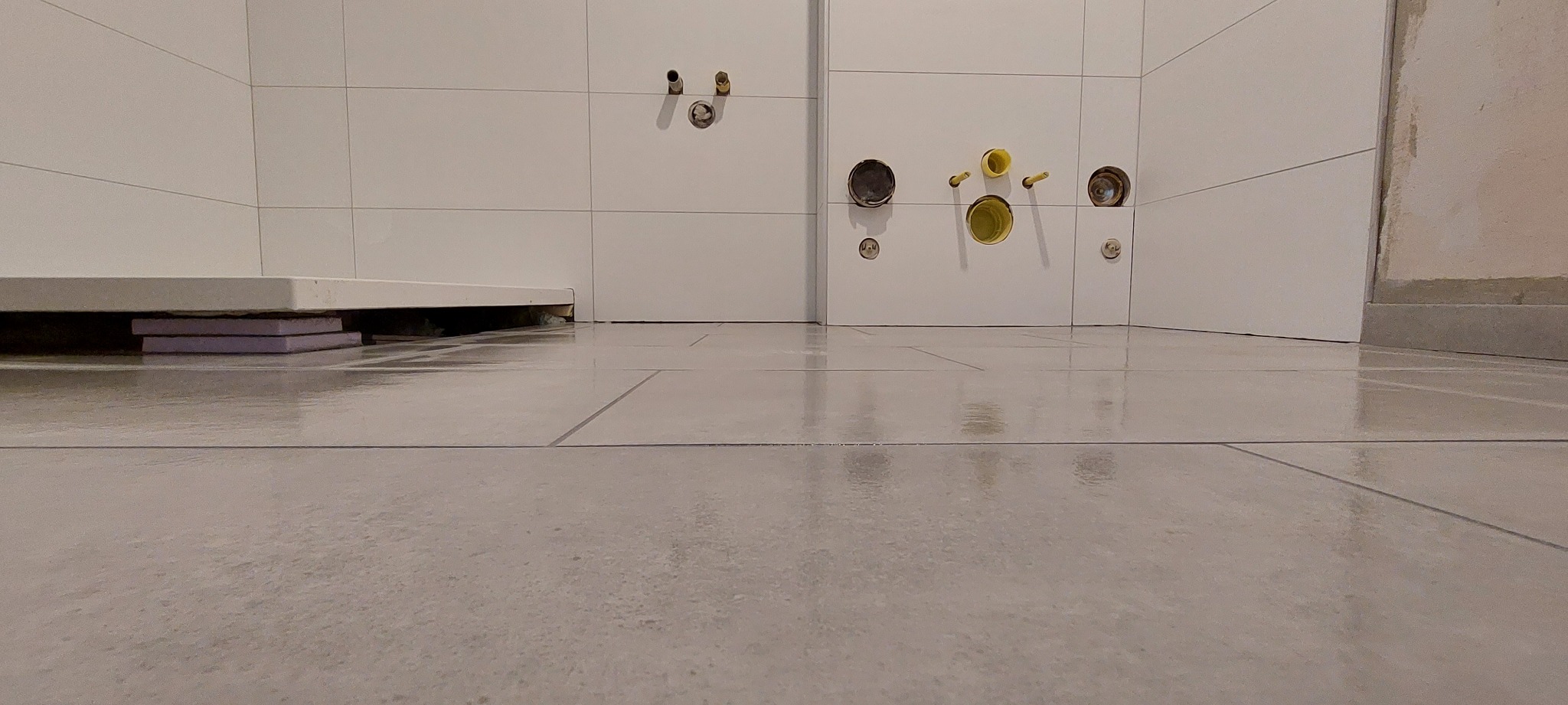 Płytki podłogowe w trakcie układania, widoczne wystające rury instalacji wodnej i kanalizacyjnej w ścianie, oraz fragment brodzika podpartego styropianem.