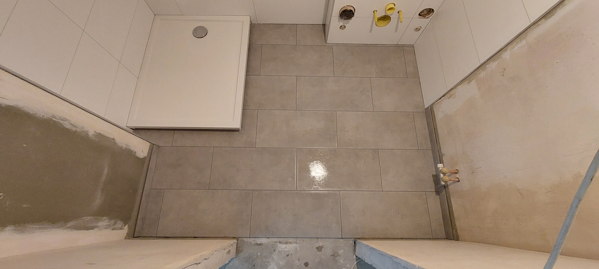 Niedokończona łazienka z szarymi płytkami podłogowymi ułożonymi w cegiełkę, biały brodzik i przygotowane podejścia wodno-kanalizacyjne na ścianie.