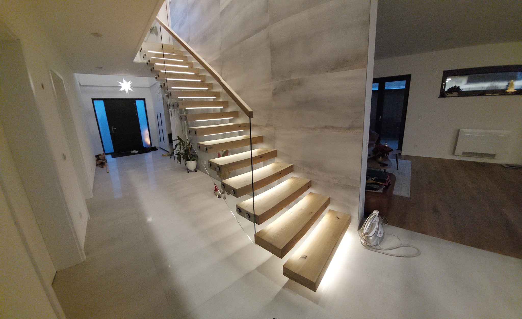 Nowoczesne schody drewniane z podświetleniem LED, zamontowane na szklanych panelach w przestronnym holu z jasną podłogą i betonową ścianą.