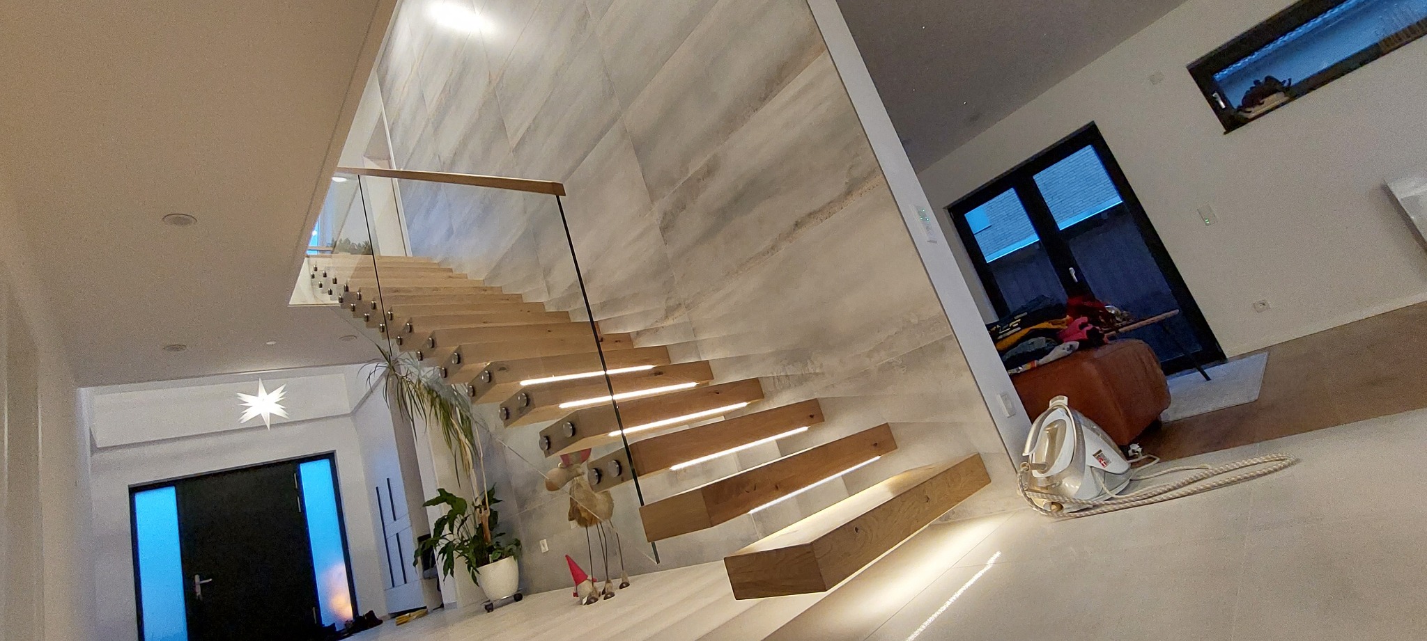 Nowoczesne schody drewniane z podświetleniem LED, szklaną balustradą i marmurową ścianą w tle, widok na korytarz z dekoracyjną gwiazdą i roślinami doniczkowymi.