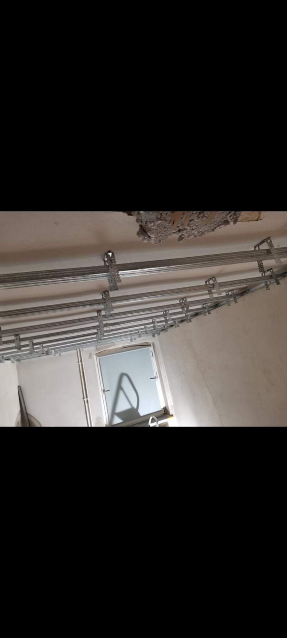 Montaż stelaża pod sufitem, widoczne profile metalowe i uchwyty mocujące, fragment uszkodzonego sufitu, okno w tle.