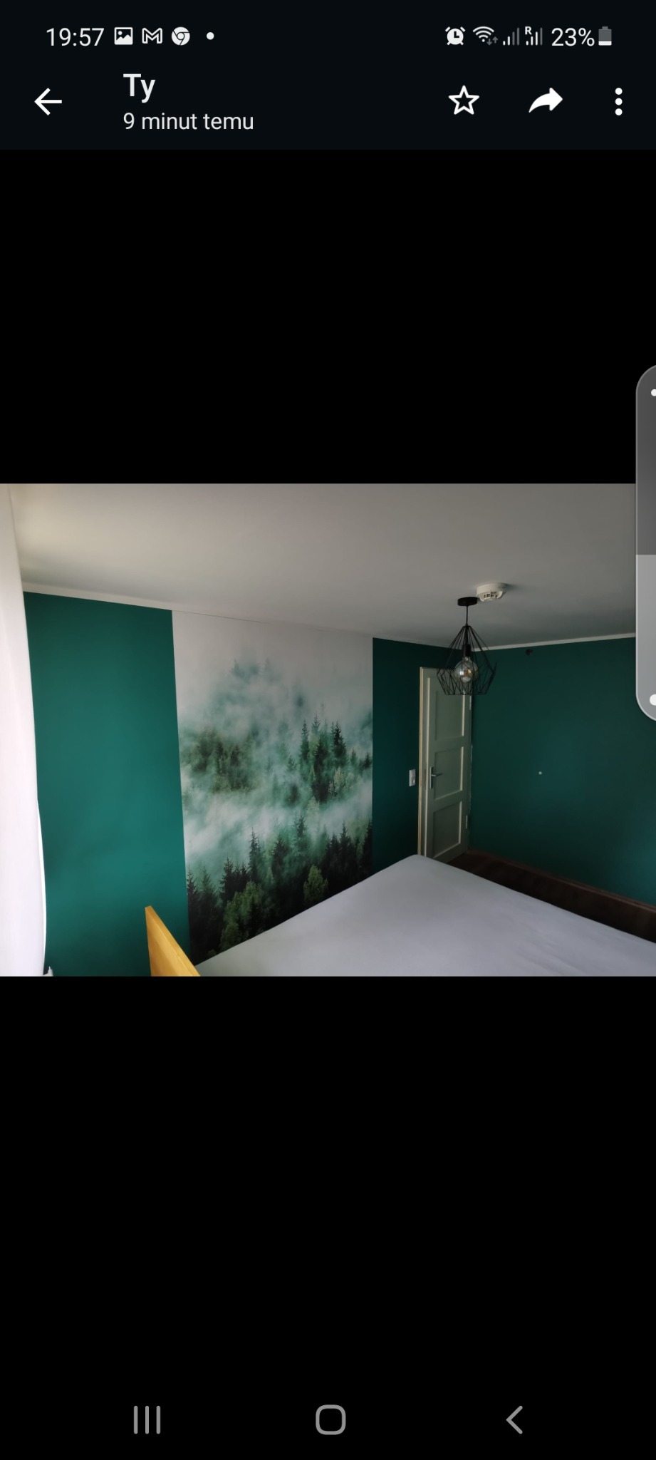 Pokój z zielonymi ścianami i fototapetą z motywem lasu we mgle, widoczna lampa wisząca w kształcie geometrycznym, fragment łóżka z białym prześcieradłem i fragment ramy łóżka z jasnego drewna.