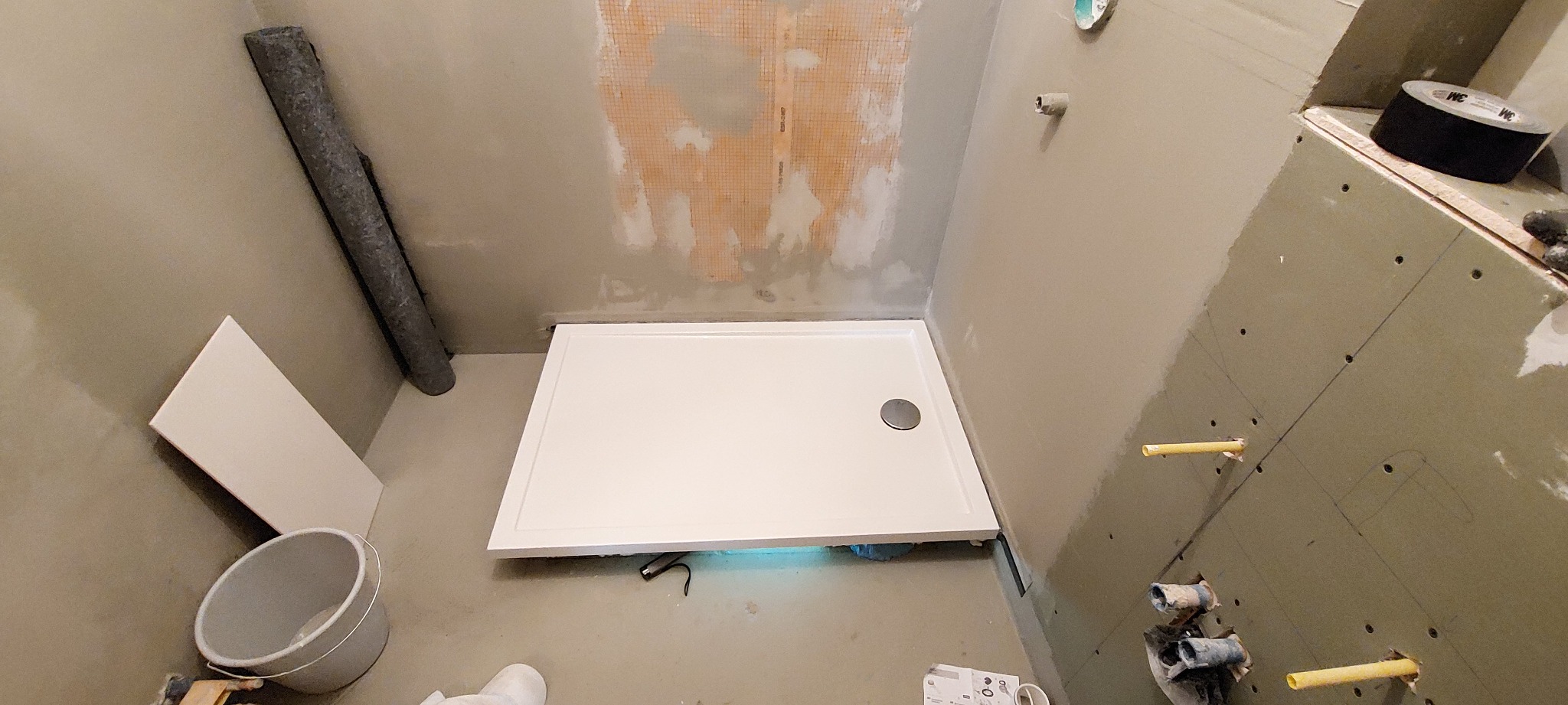 Biały brodzik w trakcie montażu w surowej łazience, widoczne przygotowane ściany z elementami instalacji wodnej, wiadro i rolka materiału izolacyjnego.