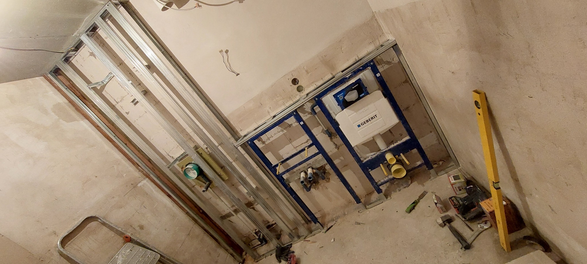 Stelaż podtynkowy Geberit zamontowany w surowej łazience, widoczne profile metalowe, instalacja wodno-kanalizacyjna, poziomica i narzędzia na podłodze.