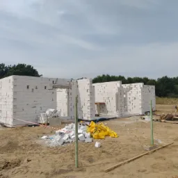 Wznoszone ściany parterowego domu z białych bloczków, widoczne otwory okienne i drzwiowe, na ziemi leżą resztki materiałów budowlanych i żółta plandeka.