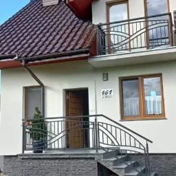 Dwupiętrowy dom z beżową elewacją, balkonem z czarną, ozdobną balustradą i schodami wejściowymi wykończonymi szarym kamieniem, z brązowym dachem i oknami.