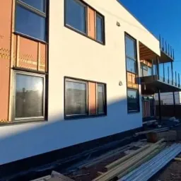 Budynek w trakcie ocieplania elewacji: widoczne fragmenty izolacji termicznej, przygotowane deski elewacyjne, okna z czarnymi ramami i jasna elewacja w słoneczny dzień.