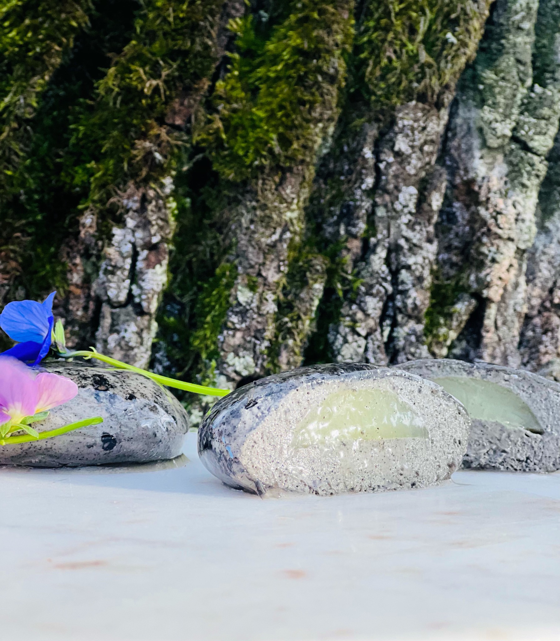 Trzy ciastka w kształcie kamieni z zielonym nadzieniem, udekorowane kwiatami na tle kory drzewa pokrytej mchem.