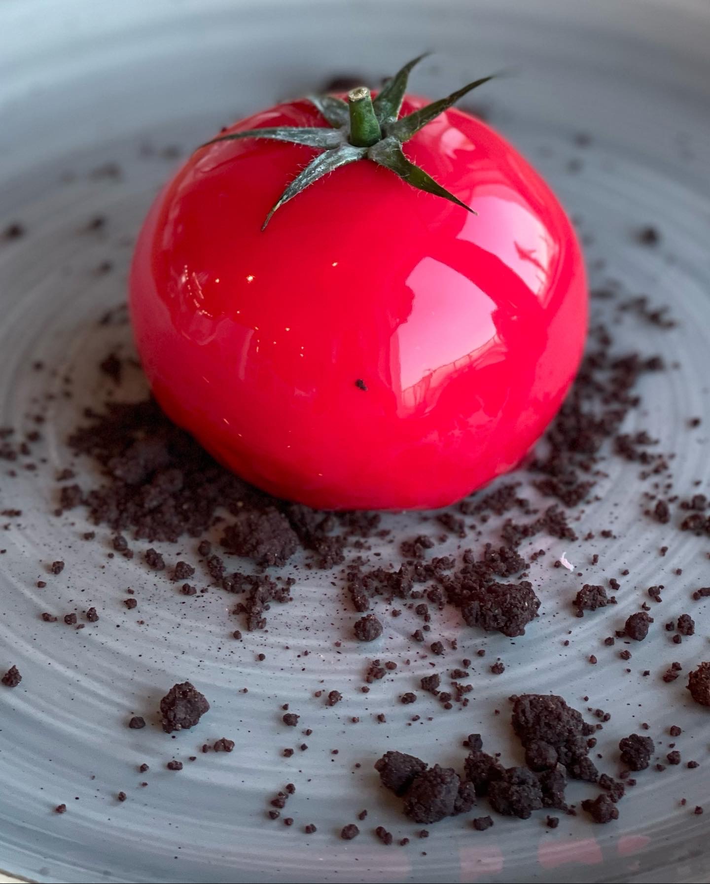 Błyszczący, czerwony deser w kształcie pomidora z zieloną szypułką, otoczony okruchami czekoladowymi na szarym talerzu.