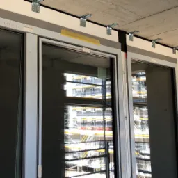 Montaż okien w surowym stanie budynku, widoczne metalowe wsporniki mocujące do sufitu, taśma malarska zabezpieczająca, budynek w tle w trakcie budowy.