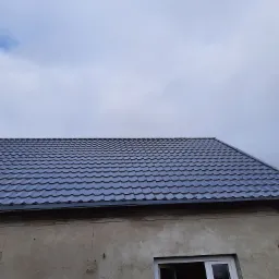 Nowy, błyszczący, grafitowy dach z blachodachówki na budynku w Gryficach, widoczny fragment ściany z oknem i pochmurne niebo.