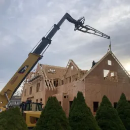 Montaż konstrukcji dachu z wykorzystaniem podnośnika teleskopowego CAT na tle wznoszonego domu w stanie surowym otoczonego zielonymi krzewami.