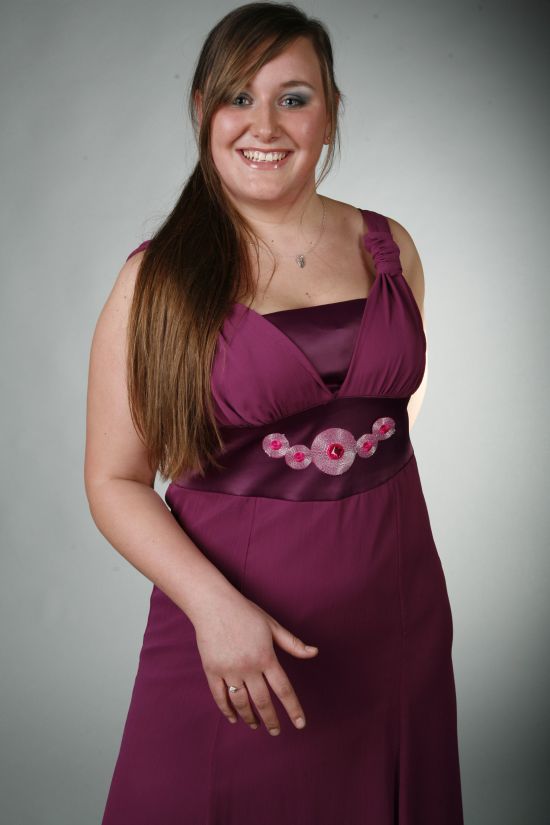 Uśmiechnięta kobieta w fioletowej sukience wizytowej z ozdobnym pasem, z długimi brązowymi włosami zaczesanymi na bok i delikatnym naszyjnikiem.