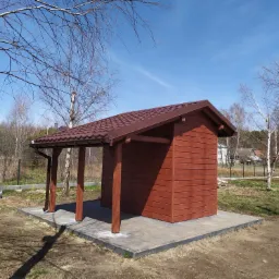 Drewniana altana z brązowym dachem krytym dachówką, zbudowana w Łańcucie, z widocznymi elementami konstrukcyjnymi i rynną, otoczona drzewami bez liści.