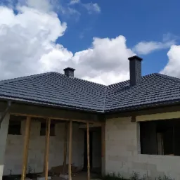 Dom w trakcie budowy z nowym, grafitowym pokryciem dachowym i dwoma kominami na tle błękitnego nieba z białymi chmurami.