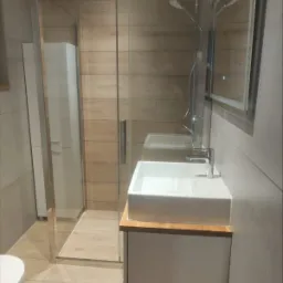 Nowoczesna łazienka z kabiną prysznicową typu walk-in, umywalką nablatową na drewnianym blacie i szarą glazurą imitującą beton.