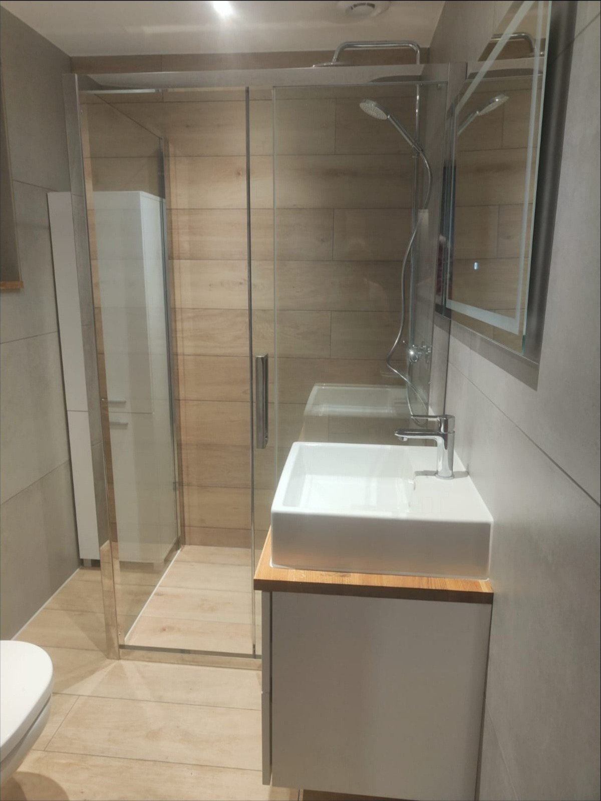 Nowoczesna łazienka z kabiną prysznicową typu walk-in, umywalką nablatową na drewnianym blacie i szarą glazurą imitującą beton.