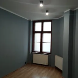 Pomalowane na szaro wnętrze pokoju z drewnianą podłogą, oknem z drewnianą ramą i dwoma grzejnikami.