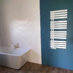 Nowoczesna łazienka z wolnostojącą białą wanną, białymi płytkami na ścianie i grzejnikiem drabinkowym na niebieskiej ścianie.