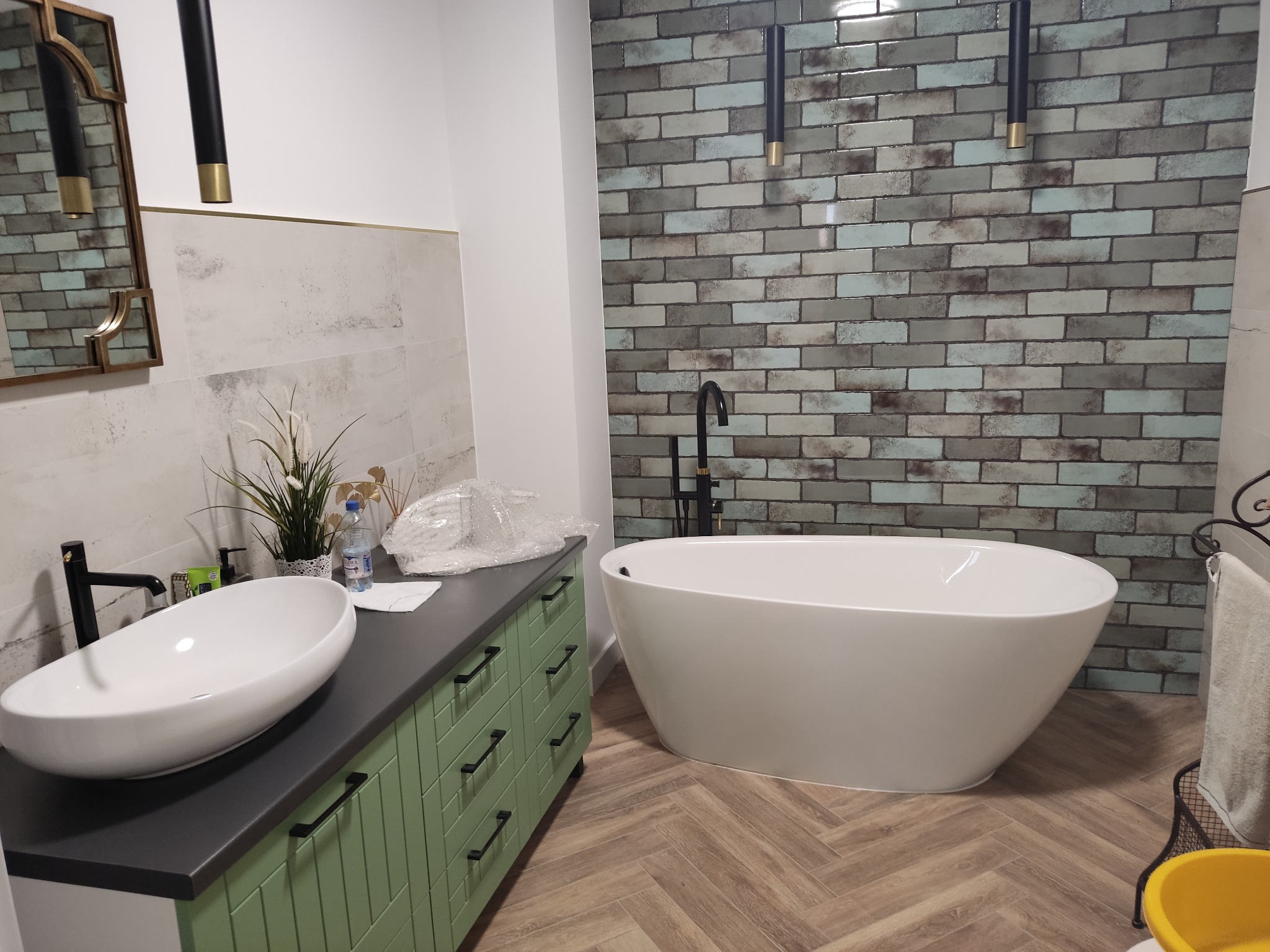 Nowoczesna łazienka z wolnostojącą wanną, umywalką nablatową na zielonej szafce z czarnym blatem oraz ścianą wyłożoną płytkami imitującymi cegłę w odcieniach szarości i błękitu.
