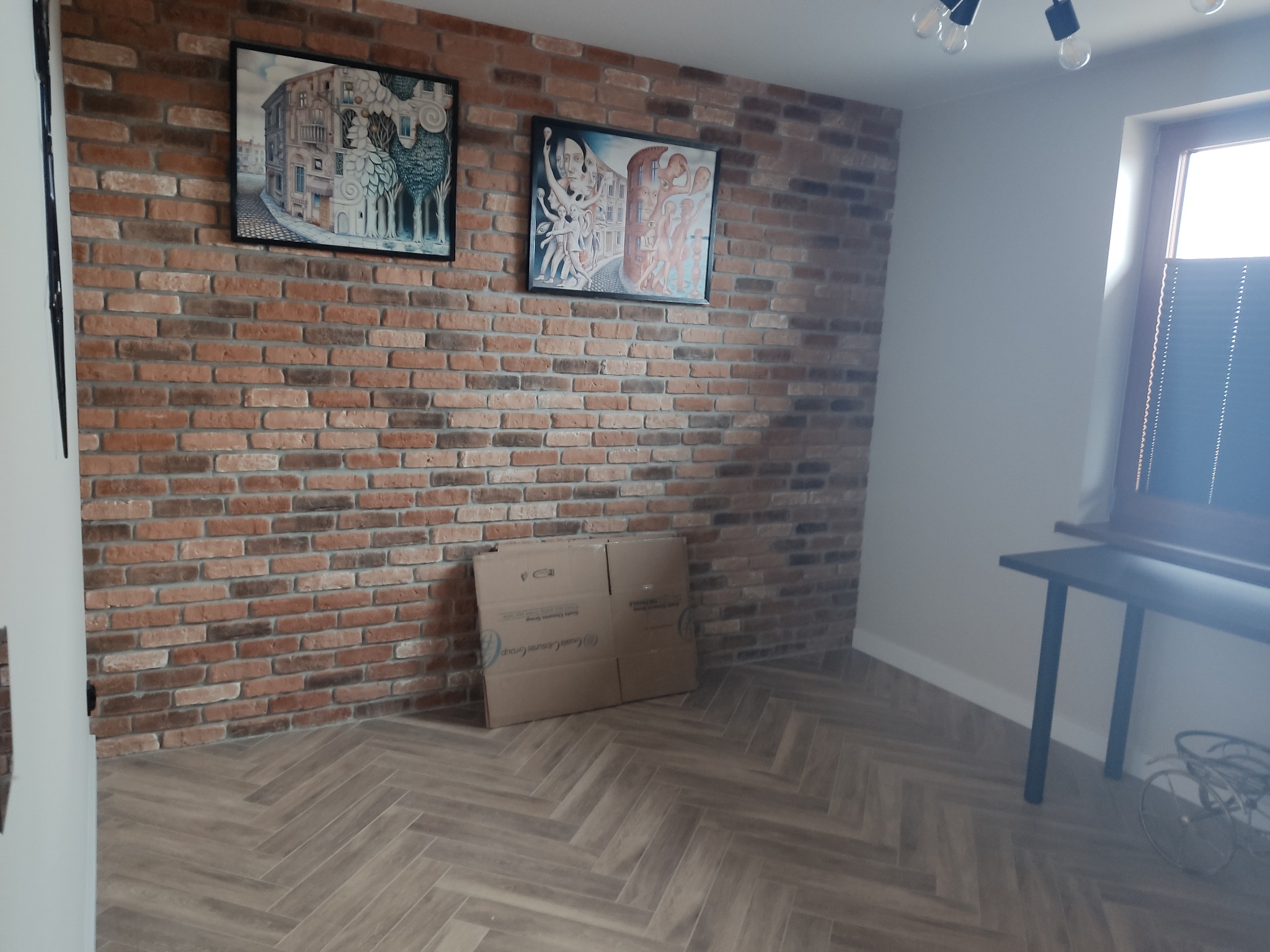Wnętrze z ceglaną ścianą, dwoma obrazami w czarnych ramkach, parkietem ułożonym we wzór jodełki oraz kartonami pod ścianą.