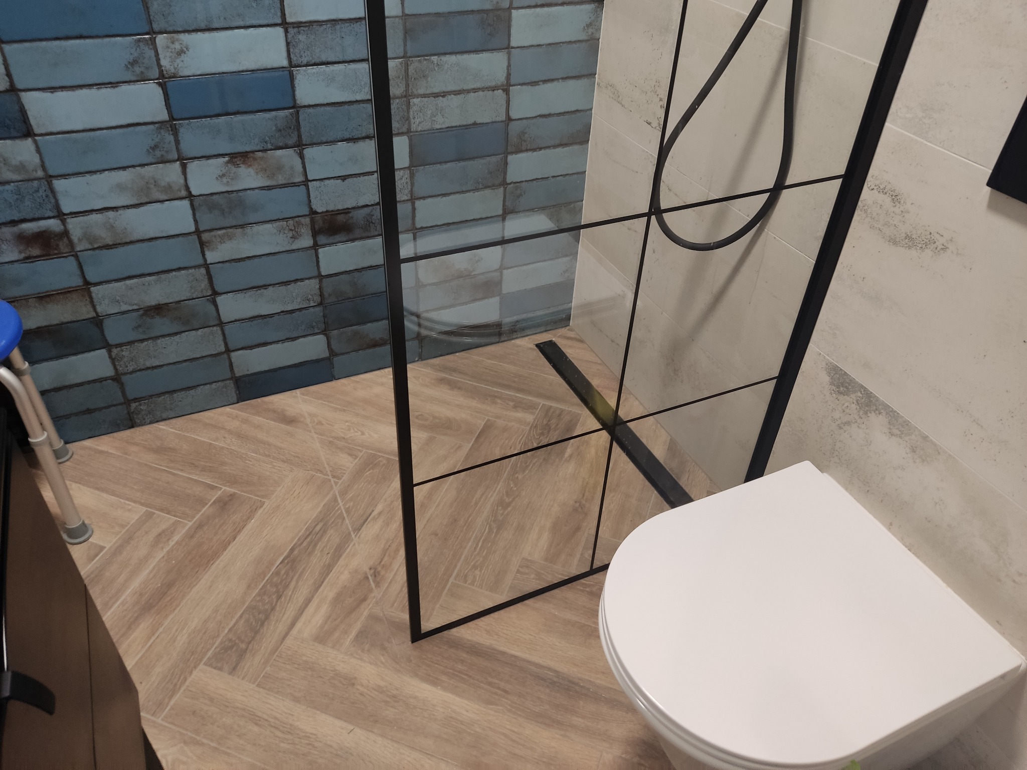 Wykończona łazienka z kabiną prysznicową typu walk-in z czarnymi profilami, toaletą wiszącą i niebieską, cegiełkową glazurą na ścianie oraz podłogą ułożoną w jodełkę.