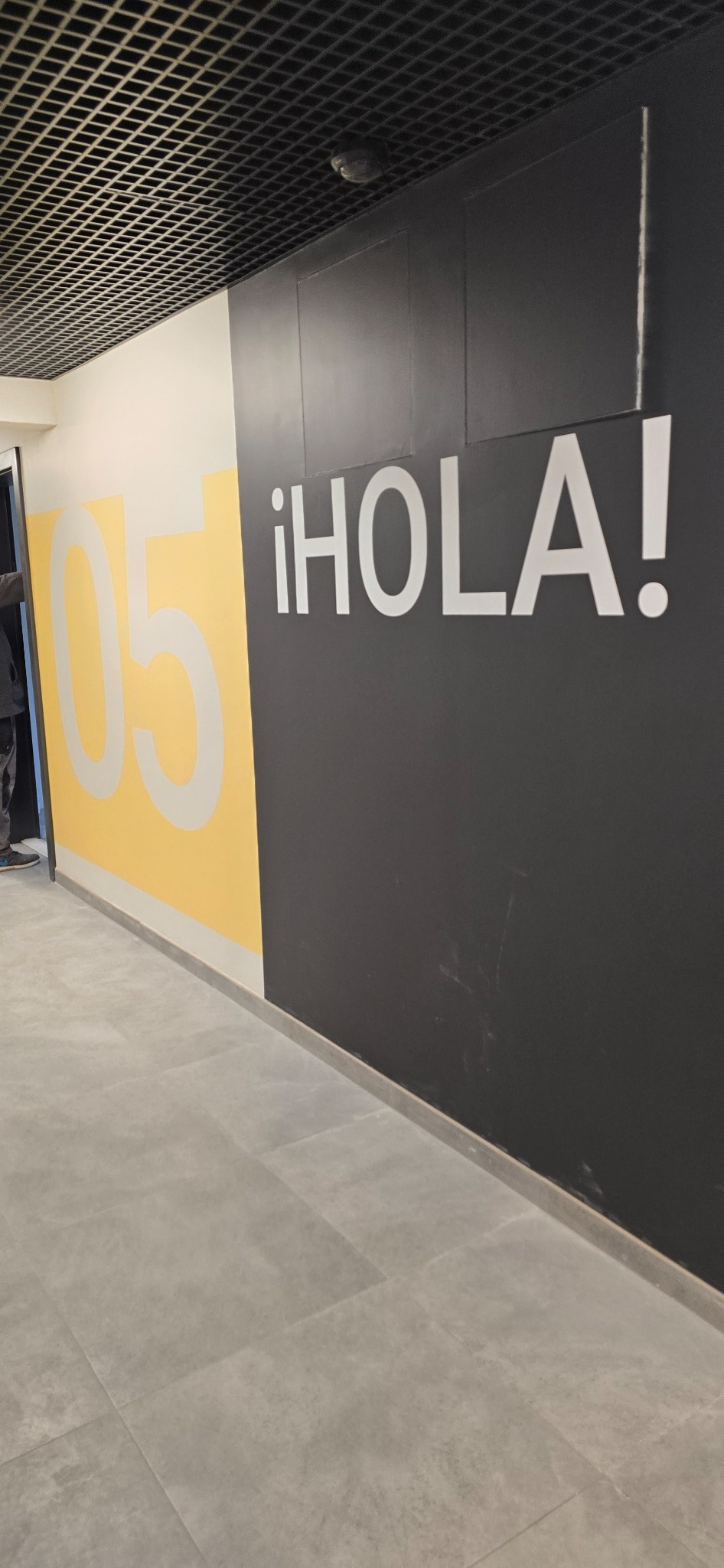 Ściana z napisem '¡HOLA!' i numerem '05' w nowoczesnym biurze. Kontrastowe kolory: czarny, biały i żółty. Minimalistyczny design i sufit rastrowy.