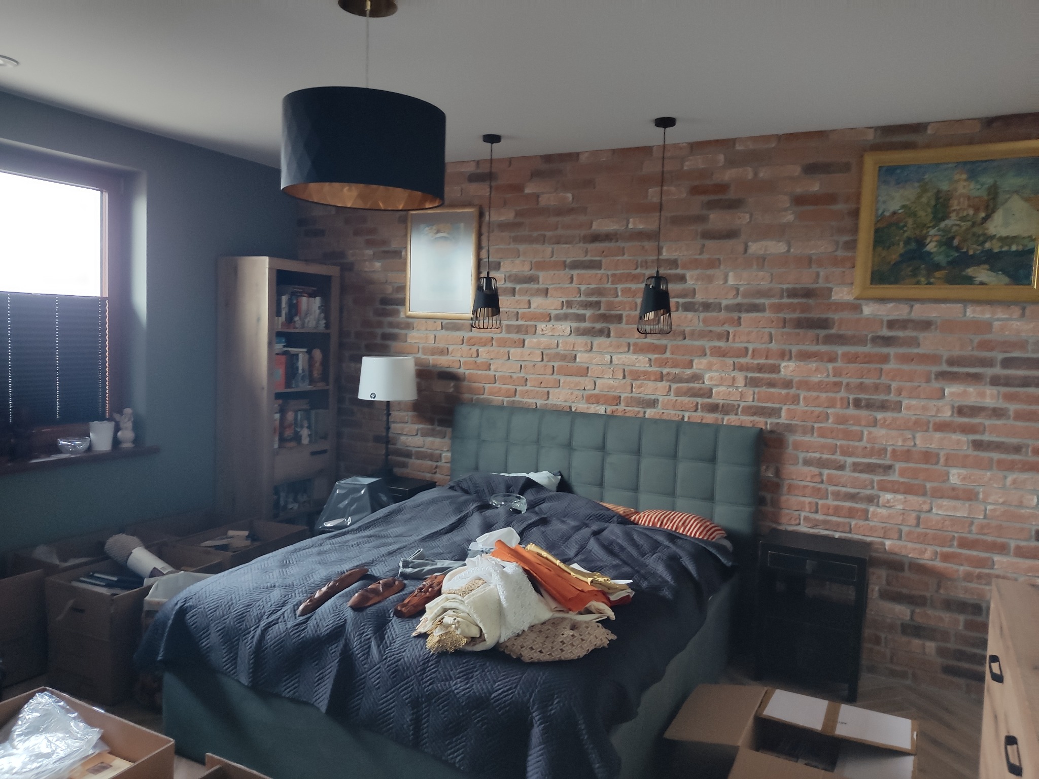 Pokój z łóżkiem, ścianą z cegły, lampami wiszącymi i kartonami, prawdopodobnie w trakcie przeprowadzki lub remontu.