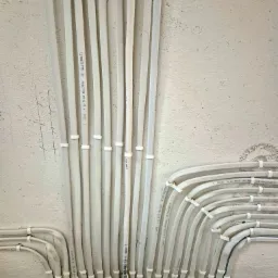 Białe kable elektryczne prowadzone po powierzchni ściany, zamocowane uchwytami. Widoczne napisy na kablach i fragment urządzenia elektrycznego IDEAL.