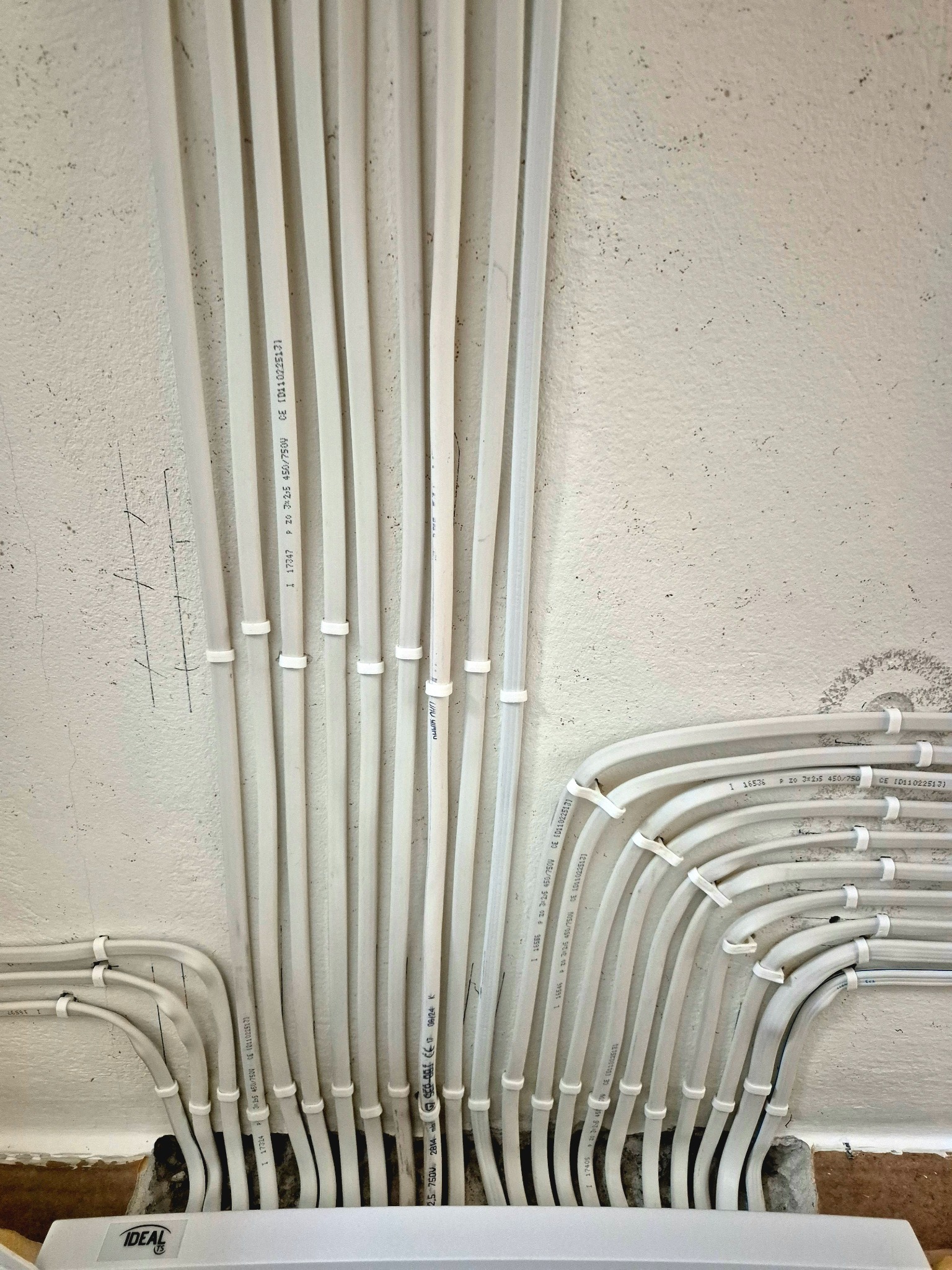 Białe kable elektryczne prowadzone po powierzchni ściany, zamocowane uchwytami. Widoczne napisy na kablach i fragment urządzenia elektrycznego IDEAL.