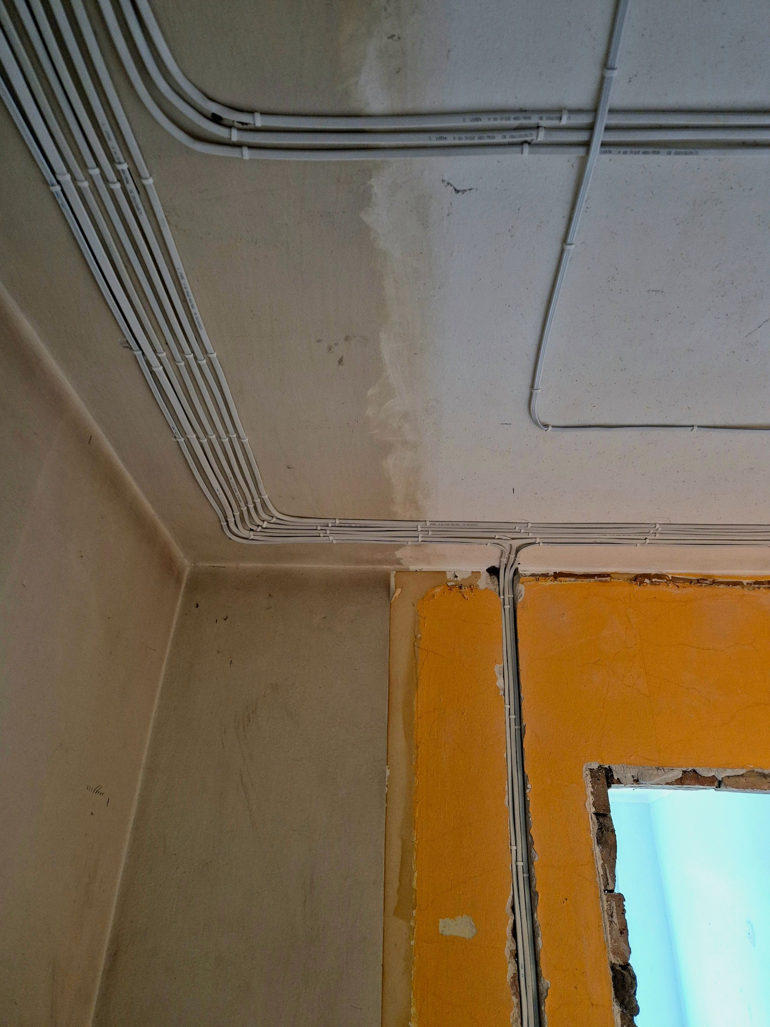 Białe, prowadzone kable elektryczne w peszlach na ścianie i suficie w surowym wnętrzu. Widoczne fragmenty ściany w kolorze żółtym i okno.