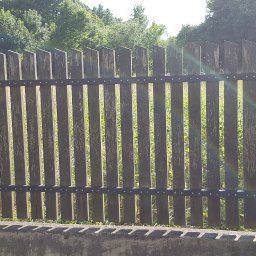 Usługi Remontowe Marcin Frączak - Drewniana, pionowa sztachetowa brama z elementami metalowymi, osadzona między dwoma murowanymi słupkami z jasnej cegły, na betonowym fundamencie, w tle zieleń drzew.