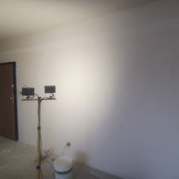 Usługi Remontowe Marcin Frączak - Wnętrze w trakcie malowania: ściany w świeżej, jasnej farbie, przenośny reflektor oświetla powierzchnię, widoczny kubeł z farbą i zabezpieczone taśmą drzwi.