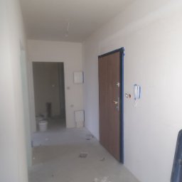 Usługi Remontowe Marcin Frączak - Korytarz w trakcie prac wykończeniowych, widoczne ślady po szpachlowaniu na podłodze i ścianach, zabezpieczone futryny drzwi wejściowych.