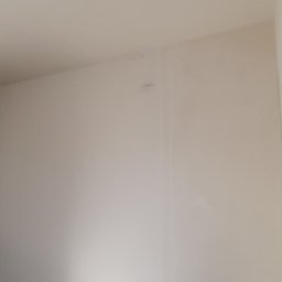 Usługi Remontowe Marcin Frączak - Świeżo pomalowane, jasne wnętrze z widocznym narożnikiem i fragmentem ościeżnicy drzwiowej. Ściana gładka, jednolita kolorystycznie, z delikatnym odblaskiem światła.