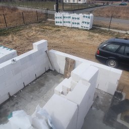 Usługi Remontowe Marcin Frączak - Wznoszenie ścian parteru z bloczków betonu komórkowego na placu budowy, widoczne palety z zapasem materiału H+H, czarny samochód kombi i motocykl w tle.