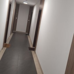 Usługi Remontowe Marcin Frączak - Korytarz w budynku z szarymi płytkami na podłodze, białymi ścianami i drewnianymi obramowaniami drzwi, oświetlony okrągłymi lampami sufitowymi.