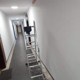 Usługi Remontowe Marcin Frączak - Korytarz w trakcie przygotowania do malowania: na ścianie widoczne pozostałości po szpachlowaniu, pod ścianą taśma malarska i resztki starej farby, rozstawiona aluminiowa drabina. Na suficie ślady...
