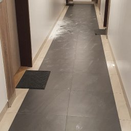 Usługi Remontowe Marcin Frączak - Długi korytarz po remoncie w Leoncinie, z ciemnymi płytkami podłogowymi pokrytymi śladami po czyszczeniu i trzema czarnymi wycieraczkami przy drzwiach.