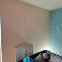 Usługi Remontowe Marcin Frączak - Przygotowanie ścian do malowania: ściana w kolorze brzoskwiniowym i błękitnym z widocznymi łatami szpachli, grzejnik pod oknem, fragment sofy z poduszkami w rogu pokoju.