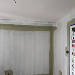 Usługi Remontowe Marcin Frączak - Wnętrze pokoju w trakcie malowania, widoczne ślady farby na ścianach i suficie, drabina malarska, drzwi oklejone naklejkami kibicowskimi.