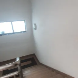 Klatka schodowa z beżowymi płytkami, białą ścianą i metalową poręczą, widok z góry, małe okno w tle.