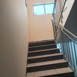 Usługi Remontowe Marcin Frączak - Klatka schodowa z wykafelkowanymi schodami w odcieniach brązu i beżu, widok z dołu, z metalową balustradą i oknem w tle, ściany pomalowane na jasny beż.