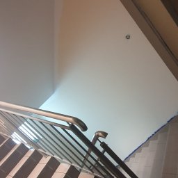 Usługi Remontowe Marcin Frączak - Schody wyłożone płytkami w czarno-beżowe pasy, z metalową balustradą, widok z dołu w kierunku sufitu z widocznym punktem oświetleniowym.