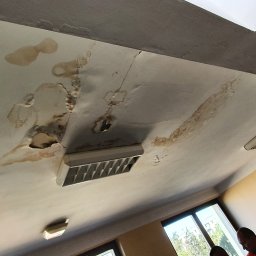Usługi Remontowe Marcin Frączak - Zacieki i uszkodzenia na suficie podwieszanym w biurze, widoczne plamy i dziury wokół kratki wentylacyjnej, w tle okno i fragment osoby w kamizelce odblaskowej.