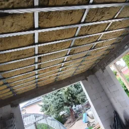 Poddasze w trakcie ocieplania, widoczne metalowe profile i wełna mineralna, ściany z białego bloczka, widok na zewnątrz na drzewa i ogród.