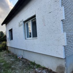 Usługi Remontowe Marcin Frączak - Ocieplanie domu styropianem: widok na ścianę budynku z nałożonymi płytami styropianowymi, częściowo jeszcze nieotynkowanymi, z widocznymi oknami i fragmentem ściany z cegły.