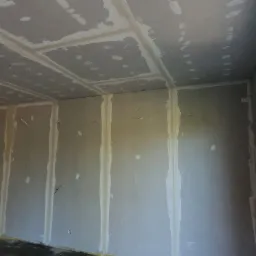 Wnętrze w trakcie wykańczania: ściany i sufit z płyt gipsowo-kartonowych, widoczne łączenia szpachlowane, okno z czerwoną izolacją, fragment ściany z ociepleniem z wełny mineralnej i bloczków...